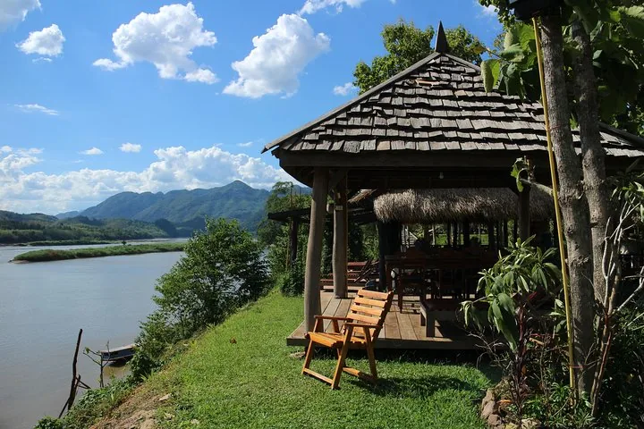 Ein hölzerner Pavillon mit Blick auf einen breiten Fluss und Berge im Hintergrund unter teilweise bewölktem Himmel.