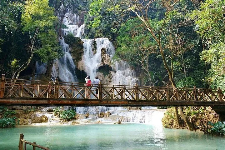 Tourists standing on a wooden bridge admiring a tall cascading waterfall in a forest.