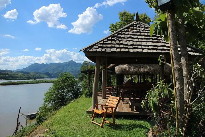 A wooden riverside pavilion with a thatched roof overlooking a wide river and mountains.