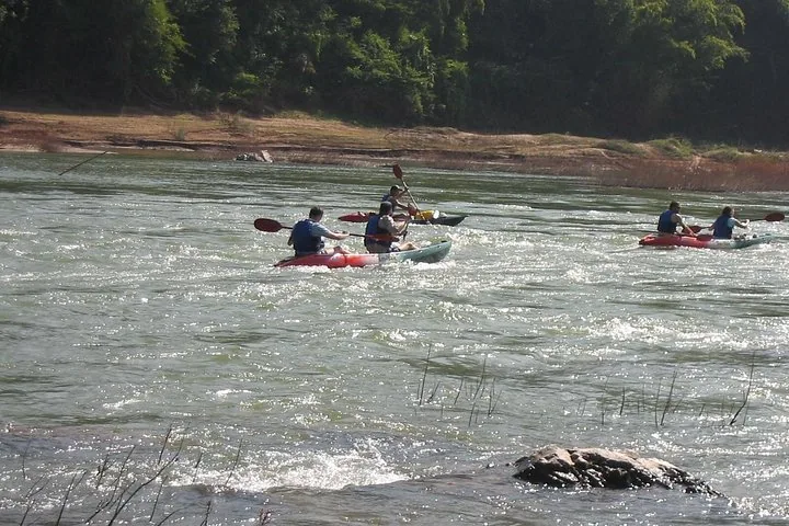 Several people paddling kayaks through small rapids on a wide river.