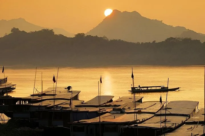 Mehrere traditionelle Flussboote liegen bei Sonnenuntergang an einem Fluss vertäut nebeneinander.
